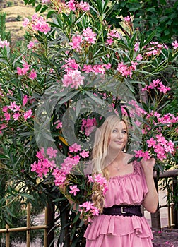 Beautiful young blonde woman in a pink dress stands posing outdoors near a bush with flowers.