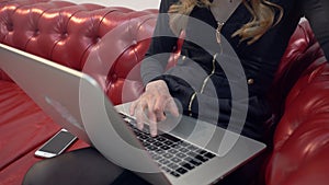 Beautiful young blonde woman on red sofa using laptop and eating a green apple in living room.