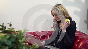 Beautiful young blonde woman on red sofa using laptop and eating a green apple in living room.