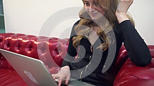 Beautiful young blonde woman on red sofa using laptop and eating a green apple in living room.