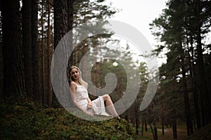 Beautiful young blonde woman sitting in forest nymph in white dress in evergreen wood