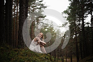 Beautiful young blonde woman sitting in forest nymph in white dress in evergreen wood