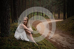 Beautiful young blonde woman sitting in forest nymph in white dress in evergreen wood