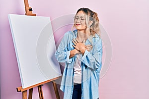 Beautiful young blonde woman standing by white painter easel stand smiling with hands on chest, eyes closed with grateful gesture