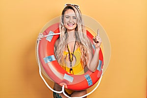 Beautiful young blonde woman wearing bikini and holding lifeguard float surprised with an idea or question pointing finger with