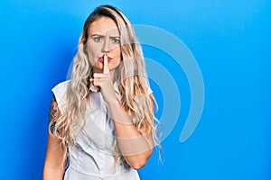 Beautiful young blonde woman wearing casual white shirt asking to be quiet with finger on lips