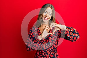 Beautiful young blonde woman wearing elegant dress smiling in love doing heart symbol shape with hands