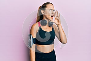 Beautiful young blonde woman wearing gym clothes and using headphones shouting and screaming loud to side with hand on mouth