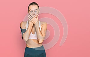 Beautiful young blonde woman wearing sportswear and arm band shocked covering mouth with hands for mistake