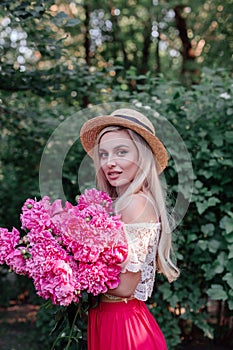 Beautiful young blonde woman wearing summer dress and straw hat standing in park posing with flower bouquet