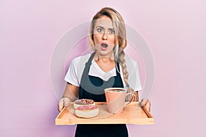 Beautiful young blonde woman wearing waitress apron holding breakfast tray in shock face, looking skeptical and sarcastic,