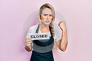 Beautiful young blonde woman wearing waitress apron holding closed banner annoyed and frustrated shouting with anger, yelling