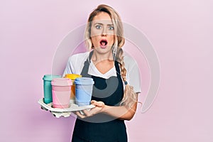 Beautiful young blonde woman wearing waitress apron holding take away cup of coffees in shock face, looking skeptical and