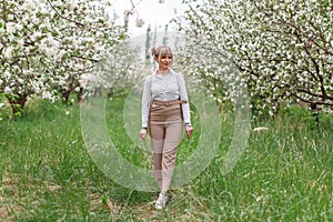 Beautiful young blonde woman in white shirt with backpack posing under apple tree in blossom in Spring garden