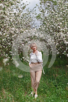 Beautiful young blonde woman in white shirt with backpack posing under apple tree in blossom in Spring garden