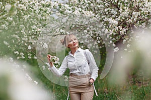 Beautiful young blonde woman in white shirt with backpack posing under apple tree in blossom in Spring garden