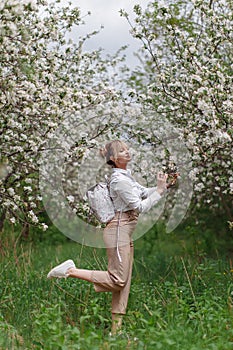 Beautiful young blonde woman in white shirt with backpack posing under apple tree in blossom in Spring garden