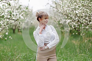 Beautiful young blonde woman in white shirt with backpack posing under apple tree in blossom in Spring garden