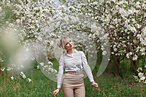 Beautiful young blonde woman in white shirt with backpack posing under apple tree in blossom in Spring garden