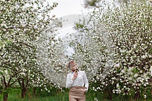 Beautiful young blonde woman in white shirt posing under apple tree in blossom in Spring garden