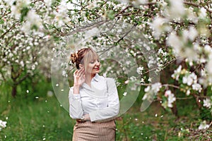 Beautiful young blonde woman in white shirt posing under apple tree in blossom in Spring garden