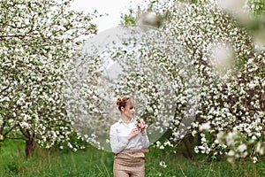 Beautiful young blonde woman in white shirt posing under apple tree in blossom in Spring garden