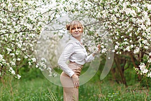 Beautiful young blonde woman in white shirt posing under apple tree in blossom in Spring garden