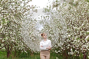 Beautiful young blonde woman in white shirt posing under apple tree in blossom in Spring garden