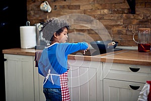 Beautiful young boy standing in a domestic kitchen by the stove, turning it on, preparing to make a meal. Young chef