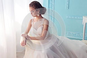 Beautiful young bride with dark curly hair in luxurious wedding dress posing at room