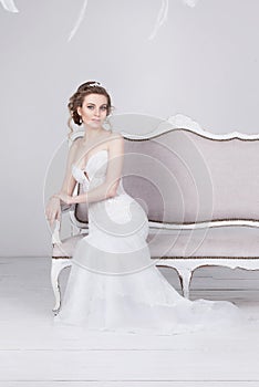 Beautiful young bride in a luxurious lace wedding dress. She sits on a white vintage sofa.