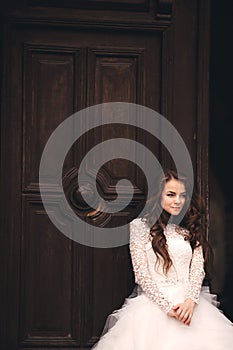 Beautiful young bride near the ancient door. Wedding portrait of a stylish woman in white long dress near old house in in a