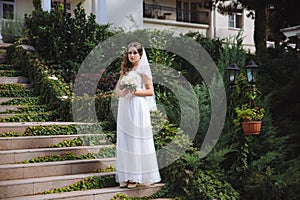 The beautiful young bride stands on stubble stairs in a dress of Greek style, her hair is curly, decorated with beads