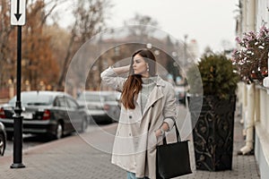 Beautiful young brown-haired woman in a sweater in a fashionable long light trench coat with a black leather bag posing on a