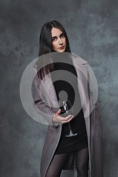 Beautiful young brunette girl in a black sweater and lilac coat on a gray background. A glass of red wine in hand.