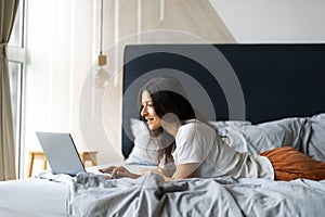 Beautiful young brunette girl with a laptop lying on the bed. Stylish modern interior. A cozy workplace. Shopping on the Internet