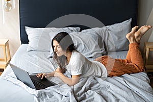 Beautiful young brunette girl with a laptop lying on the bed. Stylish modern interior. A cozy workplace. Shopping on the Internet