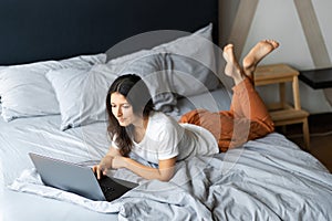 Beautiful young brunette girl with a laptop lying on the bed. Stylish modern interior. A cozy workplace. Shopping on the Internet