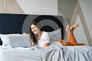 Beautiful young brunette girl with a laptop lying on the bed. Stylish modern interior. A cozy workplace. Shopping on the Internet