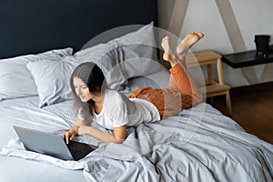 Beautiful young brunette girl with a laptop lying on the bed. Stylish modern interior. A cozy workplace. Shopping on the Internet