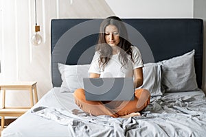 Beautiful young brunette girl with a laptop sitting on the bed. Stylish modern interior. A cozy workplace. Shopping on the