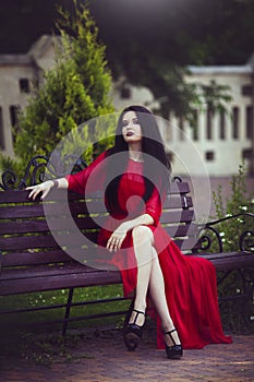 Beautiful young brunette girl in red dress is sitting on a bench.