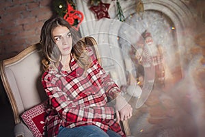 Beautiful young brunette girl in a red plaid shirt is sitting in a chair. In the photo there are Christmas lights, a fireplace, in