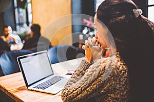 Beautiful young brunette girl in sweater working on laptop computer inside cafe at wooden table near window. Winter and Christmas