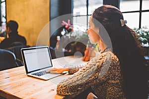 Beautiful young brunette girl in sweater working on laptop computer inside cafe at wooden table near window. Winter and Christmas