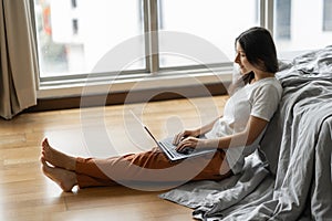 Beautiful young brunette girl working on a laptop, sitting on the floor near the bed by the panoramic window with a beautiful view