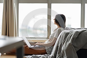 Beautiful young brunette girl working on a laptop, sitting on the floor near the bed by the panoramic window. Stylish modern