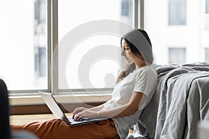 Beautiful young brunette girl working on a laptop, sitting on the floor near the bed by the panoramic window. Stylish modern