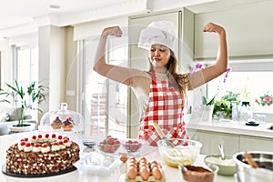 Beautiful young brunette pastry chef woman cooking pastries at the kitchen showing arms muscles smiling proud