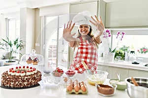 Beautiful young brunette pastry chef woman cooking pastries at the kitchen showing and pointing up with fingers number nine while